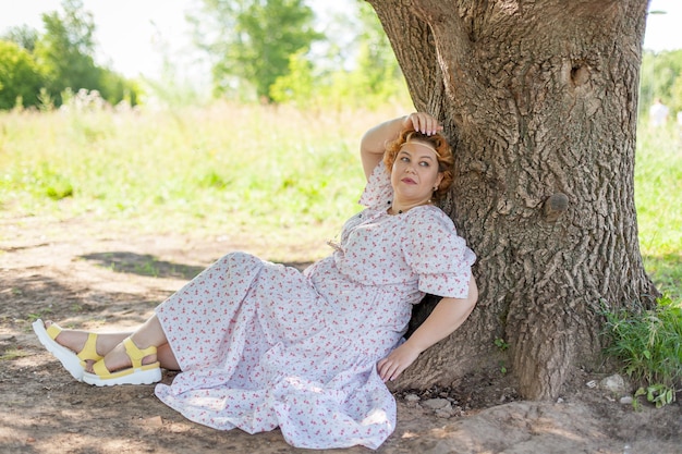 Une jeune femme vêtue d'une robe blanche, assise sur l'herbe près d'un arbre en plein air, photo d'été romantique.