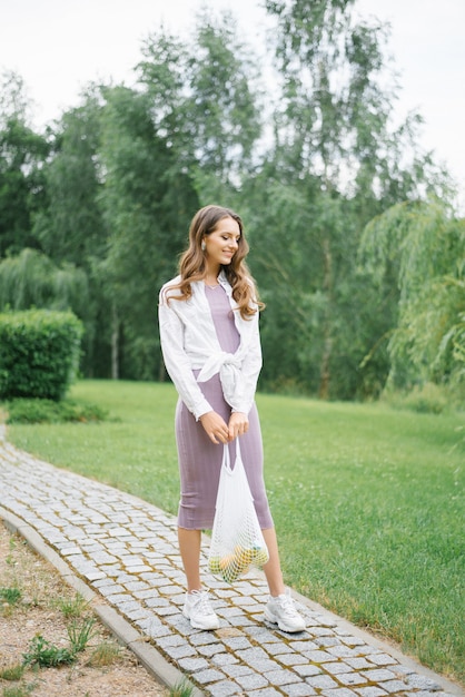 Jeune femme vêtue d'une robe lilas et d'une chemise blanche tenant un sac écologique de fruits posant dans le parc