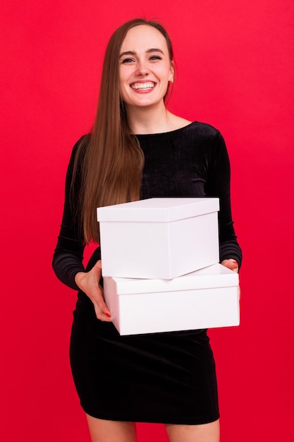 Une jeune femme vêtue d'une robe noire tient des boîtes blanches avec des cadeaux sur fond rouge Cadeaux pour les amis à Noël