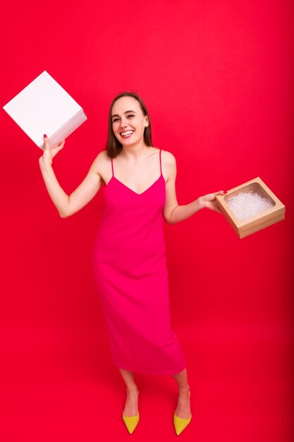 Une jeune femme vêtue d'une robe rose pose avec des cadeaux du Nouvel An sur fond rouge Photographie panoramique