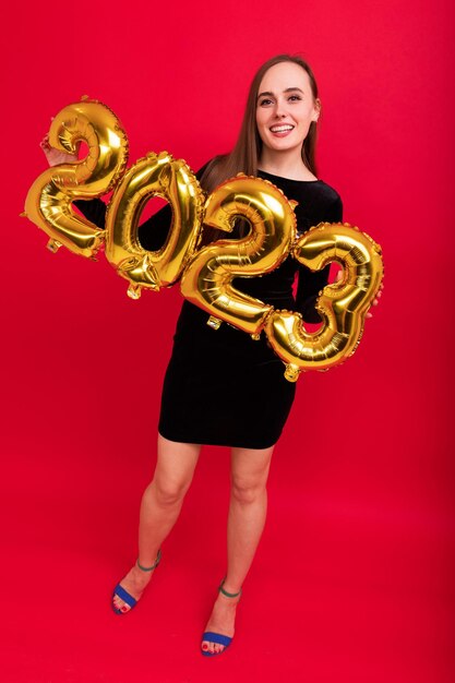 Une jeune femme vêtue d'une robe de soirée noire et de chaussures à talons hauts tient des ballons en forme de chiffres du nouvel an 2023 sur fond rouge