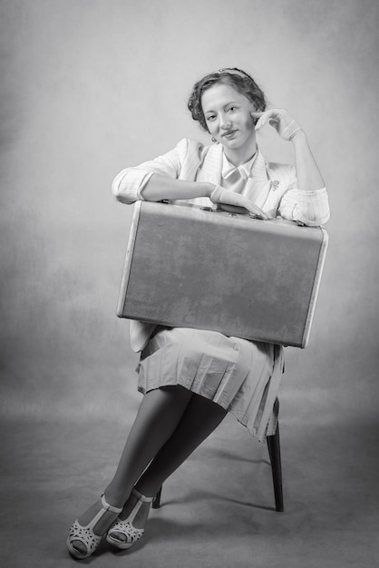 Jeune femme vêtue de style rétro assise sur une chaise avec une valise sur ses genoux