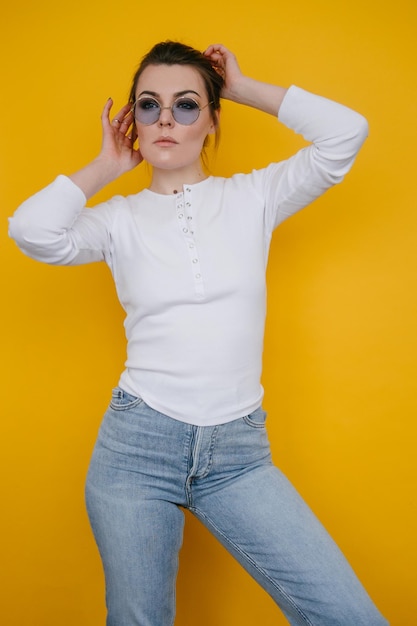 Une jeune femme vêtue d'un sweat-shirt blanc et d'un jean bleu se tient dans le studio sur un fond jaune