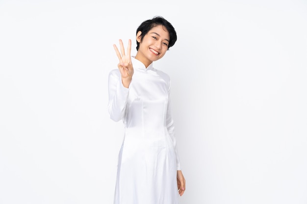 Jeune femme vietnamienne aux cheveux courts portant une robe traditionnelle sur un mur blanc heureux et en comptant trois avec les doigts