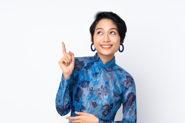 Jeune femme vietnamienne aux cheveux courts portant une robe traditionnelle sur un mur blanc isolé pointant vers le haut une excellente idée