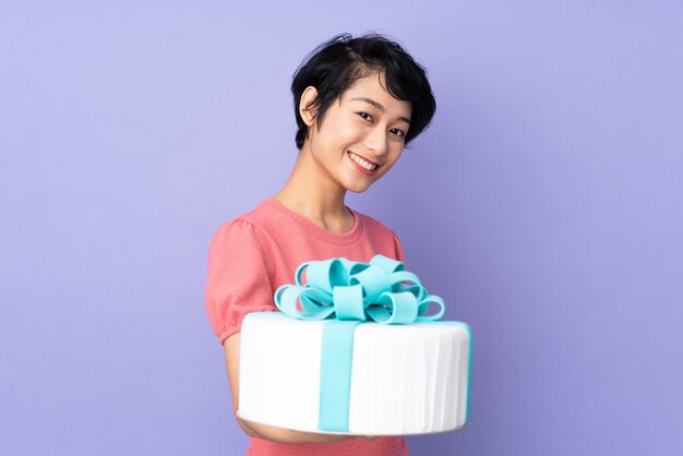 Jeune femme vietnamienne aux cheveux courts tenant un gros gâteau sur le mur violet avec une expression heureuse