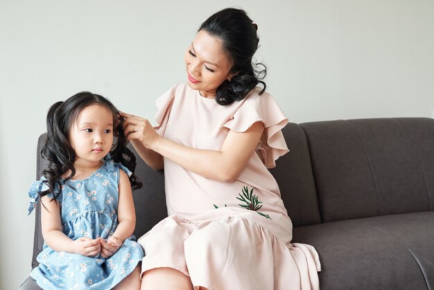 Jeune femme vietnamienne enceinte tressage les cheveux de sa petite fille sérieuse quand ils se préparent le matin