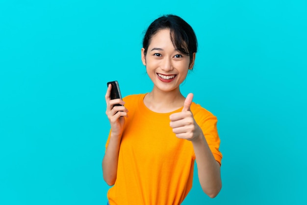 Jeune femme vietnamienne isolée sur fond bleu à l'aide d'un téléphone portable tout en faisant des pouces vers le haut