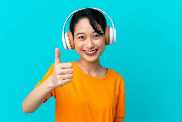 Jeune femme vietnamienne isolée sur fond bleu écoutant de la musique et avec le pouce vers le haut