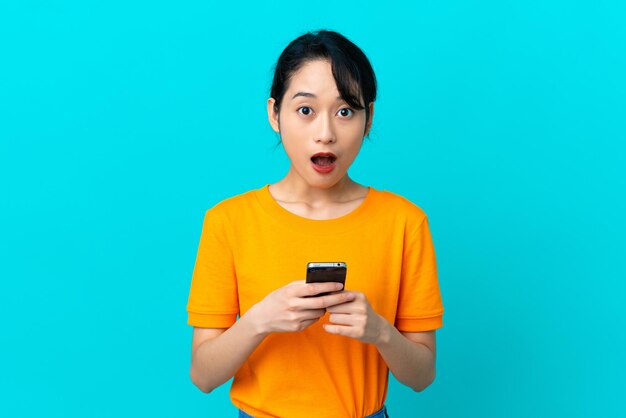 Jeune femme vietnamienne isolée sur fond bleu regardant la caméra tout en utilisant le mobile avec une expression surprise