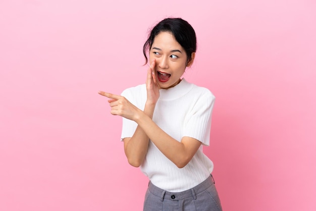 Jeune femme vietnamienne isolée sur fond rose pointant vers le côté pour présenter un produit et chuchoter quelque chose