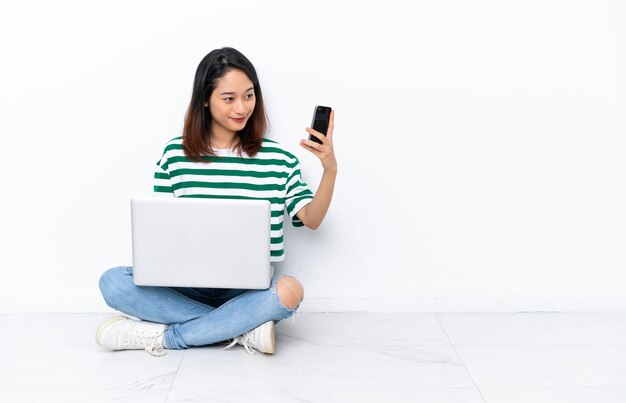 Jeune femme vietnamienne avec un ordinateur portable assis sur le sol isolé sur un mur blanc faisant un selfie