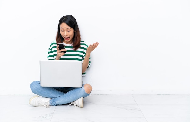 Jeune femme vietnamienne avec un ordinateur portable assis sur le sol isolé sur un mur blanc surpris et envoyant un message