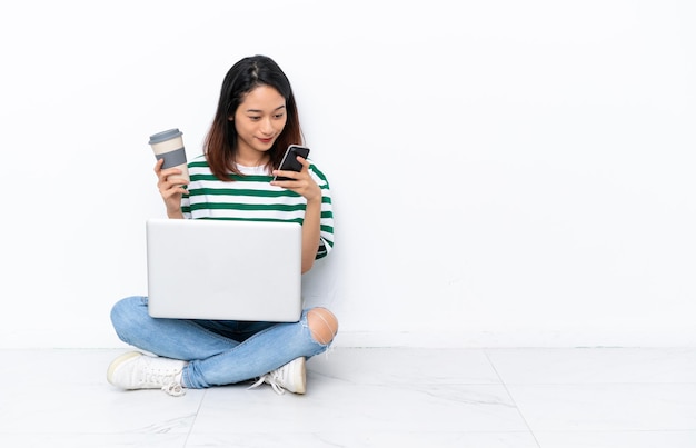 Jeune femme vietnamienne avec un ordinateur portable assis sur le sol isolé sur un mur blanc tenant du café à emporter et un mobile