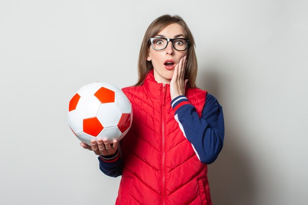 Jeune femme avec un visage surpris dans un gilet rouge tient un ballon de football dans ses mains