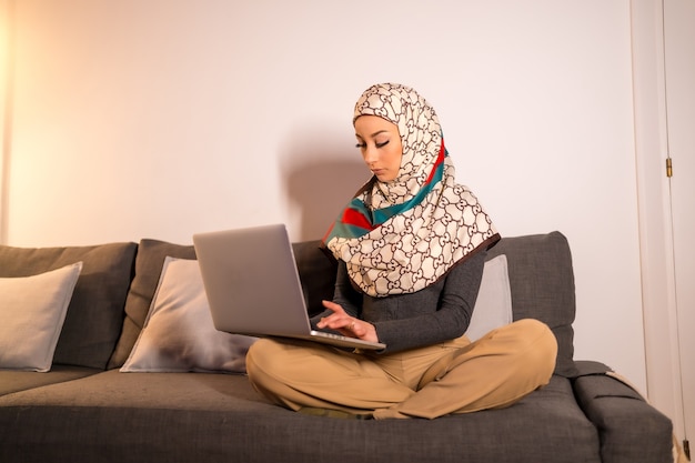 Jeune femme avec un voile blanc à son ordinateur personnel sur le canapé de son salon, faisant un travail