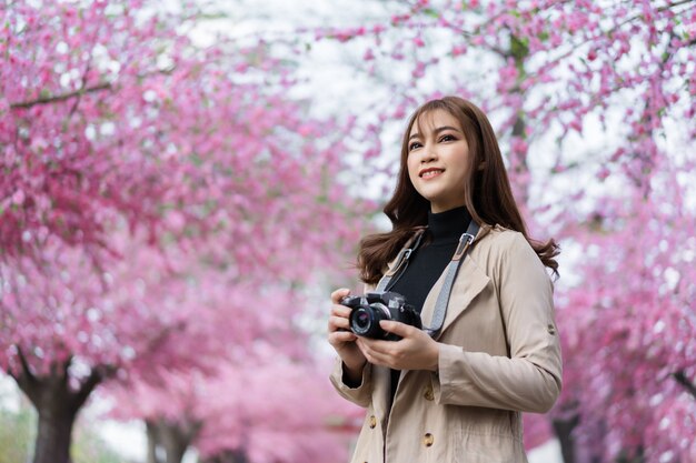 Jeune femme voyageur à la recherche de fleurs de cerisier ou de fleurs de sakura en fleurs et tenant la caméra pour prendre une photo dans le parc
