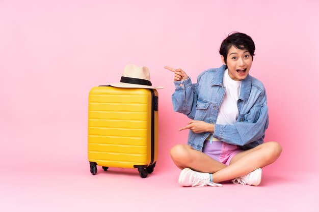 Jeune femme voyageur avec valise assis sur le sol sur le côté surpris et pointant rose