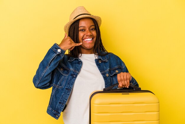 Jeune femme voyageuse afro-américaine tenant une valise isolée sur fond jaune montrant un geste d'appel de téléphone portable avec les doigts.