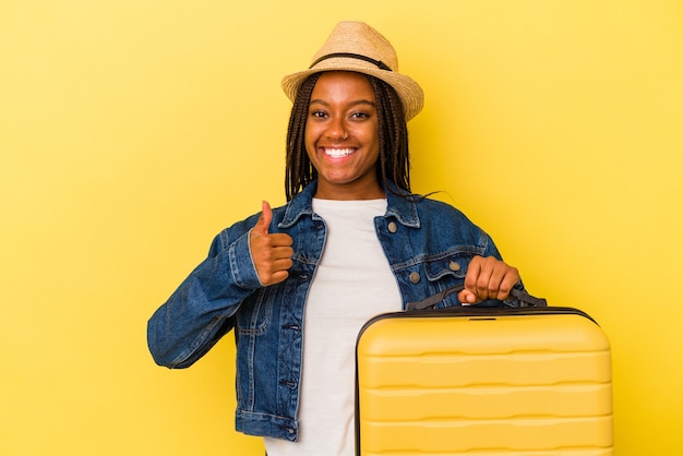 Jeune femme voyageuse afro-américaine tenant une valise isolée sur fond jaune souriant et levant le pouce vers le haut