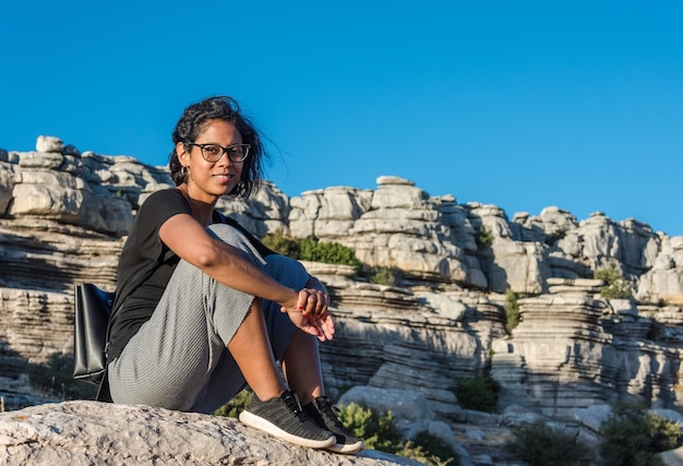 Jeune femme voyageuse latine avec des lunettes assise sur un rocher regardant directement la caméra avec un paysage de roche calcaire en arrière-plan