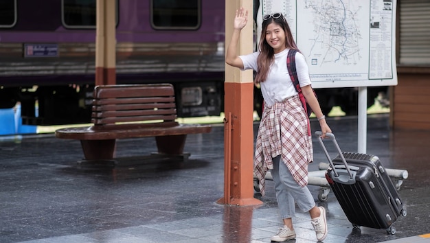 Jeune femme voyageuse millénaire agitant la main, dites bonjour, portant sa valise noire sur un arrêt de bus ou de tramway moderne vide.