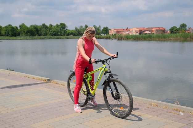 Jeune femme avec un VTT jaune sur un chemin au bord du lac Marcher à vélo dans le parc au bord de l'étang Fille avec des nattes de fils