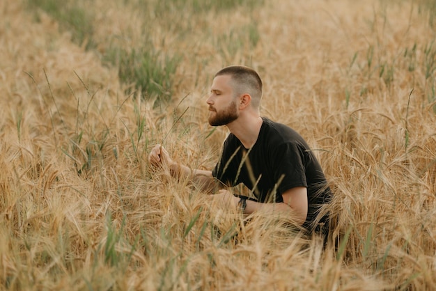 Un jeune fermier avec une barbe est assis sur le champ et teste l'épi de blé le soir.