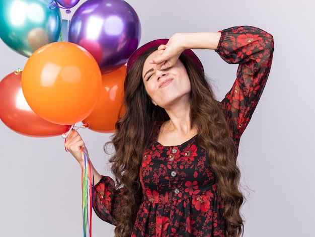 Jeune fêtarde fatiguée portant un chapeau de fête tenant des ballons en gardant le poing sur les yeux s'étirant avec les yeux fermés isolés sur un mur blanc