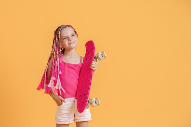 Jeune fille active avec des dreadlocks en chemise rose et short avec pennyboard sur fond jaune