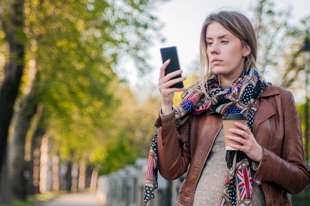 Une jeune fille adolescent de Hipster inquiète regarde son téléphone intelligent dans un parc avec un fond non focalisé