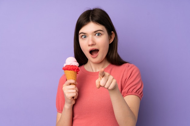 Jeune fille adolescente ukrainienne tenant une glace au cornet sur un mur violet surpris et pointant vers l'avant