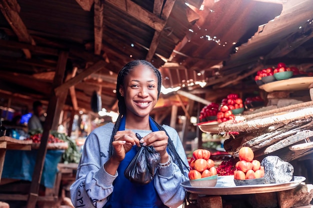 Une jeune fille africaine portant un tablier coloré vendant des tomates et des légumes dans un marché africain local typique