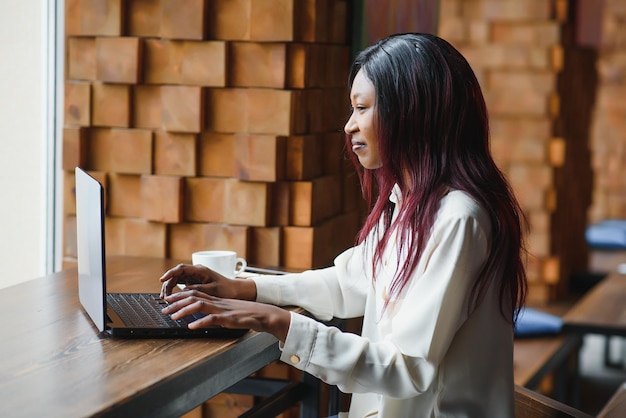 Jeune fille afro-américaine assise au restaurant et tapant sur son ordinateur portable Jolie fille travaillant sur ordinateur au café