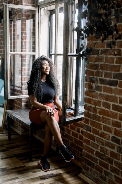 Jeune fille afro-américaine assise sur un mur de briques avec une fenêtre en t-shirt noir et pantalon rouge.