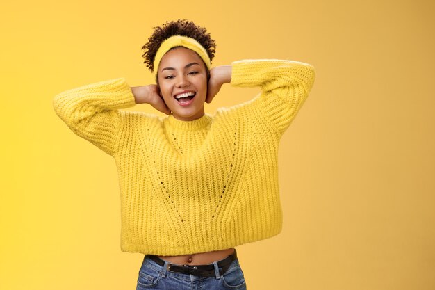 Une jeune fille afro-américaine charmante et insouciante pose les mains derrière la tête en se relaxant en s'étirant se sentir paresseux en profitant d'un jour de congé gratuit souriant largement passer des loisirs en s'amusant sur fond jaune.