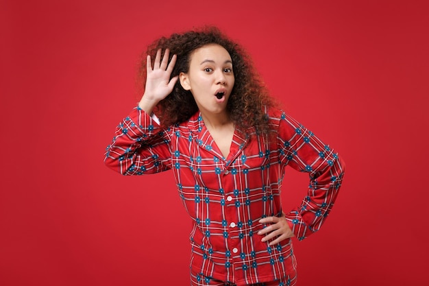 Jeune fille afro-américaine choquée en pyjama homewear posant au repos à la maison isolée sur fond rouge. Détendez-vous le concept de mode de vie de bonne humeur. Maquette de l'espace de copie. Essayez de vous entendre avec la main près de l'oreille.