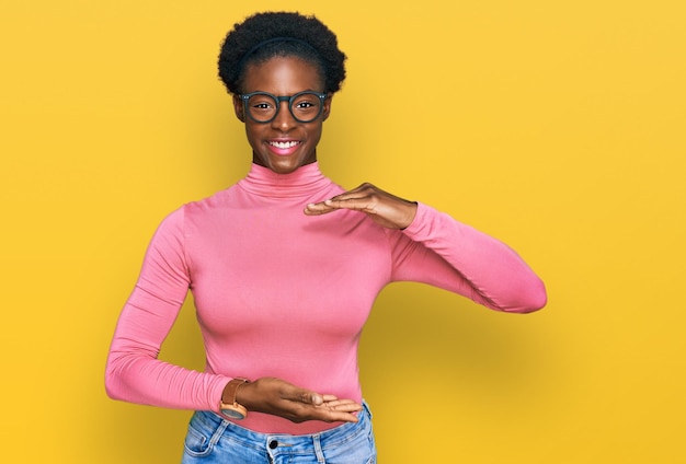 Jeune fille afro-américaine portant des vêtements décontractés et des lunettes gesticulant avec les mains montrant un signe de grande et grande taille, symbole de mesure. souriant en regardant la caméra. notion de mesure.