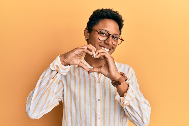 Jeune fille afro-américaine portant des vêtements décontractés et des lunettes souriant amoureux faisant la forme de symbole de coeur avec les mains concept romantique