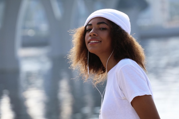 Jeune fille afro-américaine près de la mer et du pont à l'aide d'un téléphone portable et d'un casque