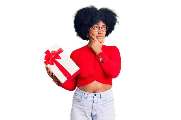 Jeune fille afro-américaine tenant un cadeau pensée inquiète au sujet d'une question préoccupée et nerveuse avec la main sur le menton
