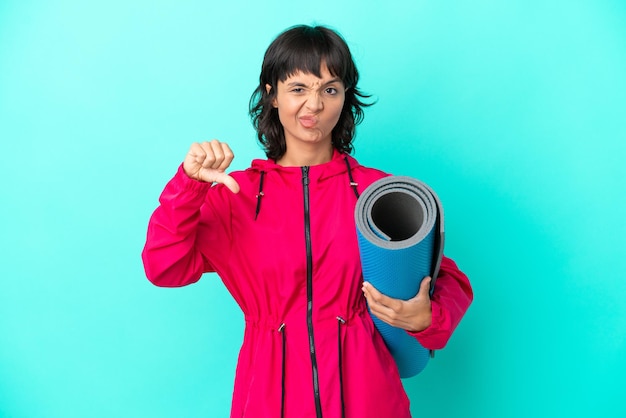 Jeune fille allant à des cours de yoga tout en tenant un tapis isolé sur fond bleu montrant le pouce vers le bas avec une expression négative