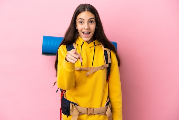 Jeune fille d'alpiniste avec un gros sac à dos isolé sur fond rose surpris et pointant vers l'avant