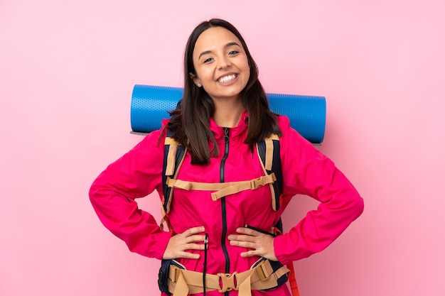 Jeune fille d'alpiniste avec un gros sac à dos sur un mur rose isolé posant avec les bras à la hanche et souriant