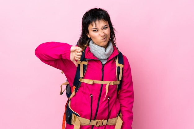 Jeune fille alpiniste avec un gros sac à dos sur le mur rose montrant le pouce vers le bas signe