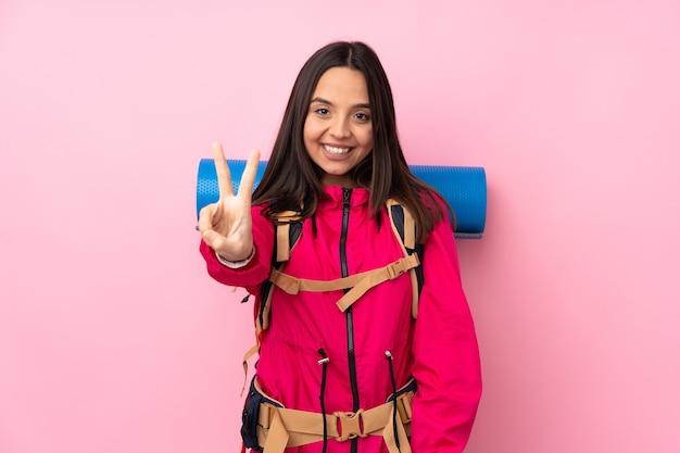 Jeune fille d'alpiniste avec un gros sac à dos sur rose isolé souriant et montrant le signe de la victoire