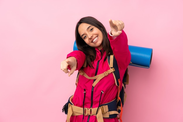 Jeune fille d'alpiniste avec un gros sac à dos sur rose pointe le doigt sur vous en souriant