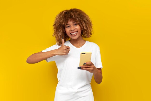 une jeune fille américaine bouclée avec des bretelles utilise un smartphone et montre comme sur fond jaune