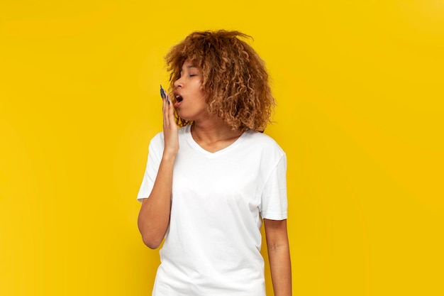 Une jeune fille américaine bouclée et fatiguée bâille et se couvre la bouche avec sa main sur un fond jaune.