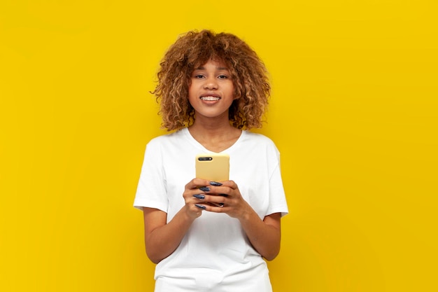 Jeune fille américaine bouclée en t-shirt blanc à bretelles utilise un smartphone sur fond jaune